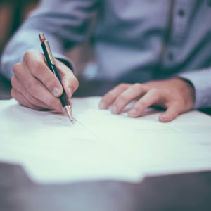 A person in a blue shirt writing on papers with a pen on a desk.